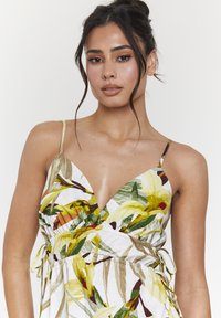 Vestido floral en blanco con hojas verdes y amarillas. Con un escote en V profundo, tirantes finos y lazos laterales para un ajuste ajustable.