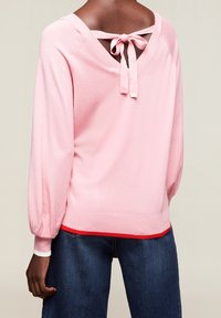 Ljust rosa tröja med V-ringning, knytdetalj på baksidan och puffärmar. Har en röd accent vid fållen. Mjuk, texturerad tyg.