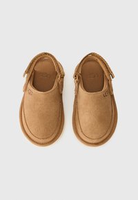 Chaussures à enfiler en suède taupe avec un bout rond, surface texturée et détails cousus. Elles possèdent une doublure intérieure douce et un logo sur le côté.