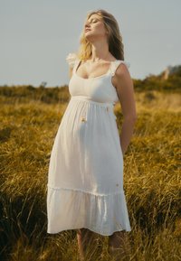 Robe maxi blanche fluide avec un ourlet volanté et des manches courtes volantées. Présente un corsage ajusté et des accents en matériel doré décoratifs.