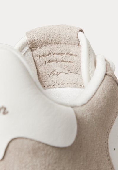Beigen nupukkinahkainen kengän kanta, jossa on valkoinen nahkainen yksityiskohta ja kauluksen sisäpuolelle painettu lainaus: "En suunnittele vaatteita. Suunnittelen unelmia."