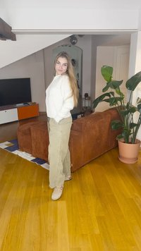 Smėlio spalvos dryžuotas korduroso sofa, šviesiai žali plataus pjūvio kelnės, balta megztinis, medinės grindys, vidaus augalas ir televizorius su spalvotu kabinetu.