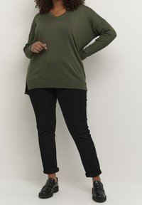 Sweter w kolorze zielonym z dekoltem w serek, o oversize'owym kroju i prążkowanej fakturze, z bocznymi rozcięciami, zestawiony z czarnymi dżinsami z podwiniętymi nogawkami oraz czarnymi mokasynami.