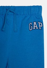 Pantalons de survêtement en coton bleu avec une taille élastique, dotés d'un cordon de serrage et d'un logo "GAP" brodé en blanc et gris sur le côté gauche.