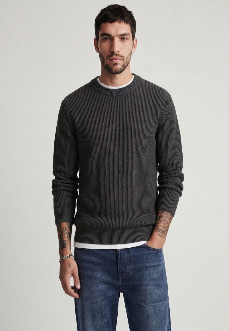 Mann mit kurzen dunklen Haaren und Tattoos trägt einen dunkelgrauen Rippsweater über einem weißen Hemd und blauen Jeans und steht vor einem schlichten Hintergrund.