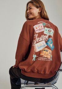 Brun sweatshirt med en afslappet pasform, der har farverige trykte chokoladeetiketter på ryggen, inklusive tekst og dekorative sløjfer.
