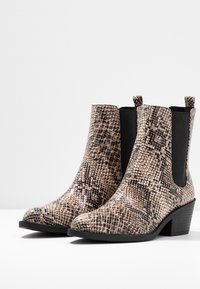 Une paire de bottines hautes au niveau de la cheville avec un motif en peau de serpent marron et noir, des pointes effilées, des panneaux latéraux en élastique et de petits talons carrés sur une surface blanche.