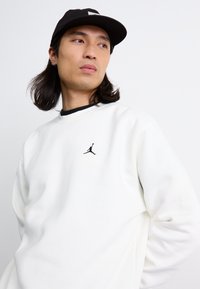 Hvid sweatshirt med rund halsudskæring, blød tekstur og sort Jordan-logo på brystet. Sammen med en sort kasket, der viser et afslappet look.