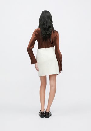 Camisa satinada marrón de manga larga con puño acampanado, combinada con una minifalda blanca. Zapatos negros con suela baja y gruesa completan el conjunto.