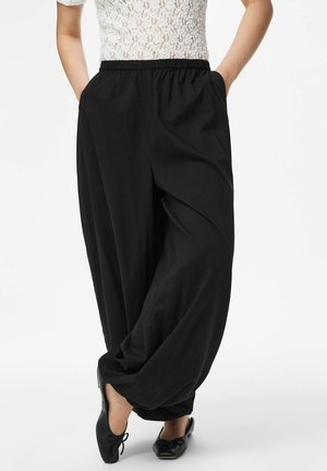 Kobieta w luźnych czarnych spodniach z elastycznym pasem i czarnych balerinach, stojąca z rękami w kieszeniach na białym tle.