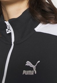 Sort lynlåsjakke med hvide detaljer, der har en opretstående krave og et sølvfarvet Puma-logo på brystet. Glat stofstruktur.