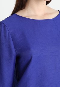 Blusa viola con una texture liscia, scollatura ampia e maniche arricciate, che mette in mostra un design semplice ed elegante.
