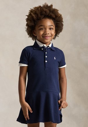 Jeune fille aux cheveux bouclés sourit, portant une robe polo bleu marine avec col et poignets blancs, sur un fond uni.
