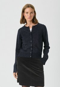 Cardigan albastru închis cu o strălucire subtilă, guler rotund, manșete cu model și închidere cu nasturi în față, asortat cu o fustă închisă.