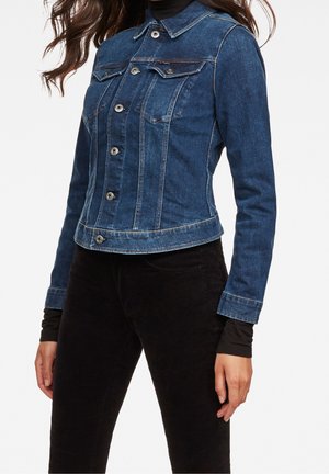 Kvinde iført en tætsiddende blå denimjakke med knaplukning over sort langærmet trøje og sorte bukser, stående mod en hvid baggrund.