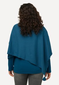 Poncho lavorato a maglia in teal con design drappeggiato, caratterizzato da un accento con frange all'orlo. Indossato sopra una maglietta a maniche lunghe e pantaloni grigi.