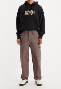 Černá mikina s kapucí a zlatým logem Levi's, spárovaná s volnými, světle hnědými cargo kalhotami s mnoha kapsami a poutek na opasek. Bílé boty.