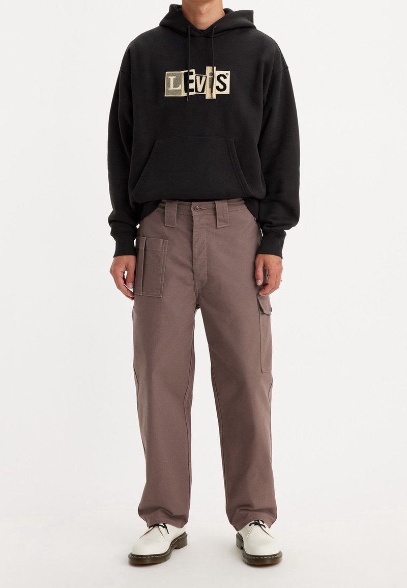Černá mikina s kapucí a zlatým logem Levi's, spárovaná s volnými, světle hnědými cargo kalhotami s mnoha kapsami a poutek na opasek. Bílé boty.
