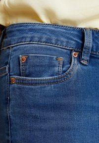 Jeans de mezclilla azul con un diseño ajustado, que cuentan con herrajes de cobre y costuras visibles. Incluyen un pequeño bolsillo frontal y trabillas para el cinturón.