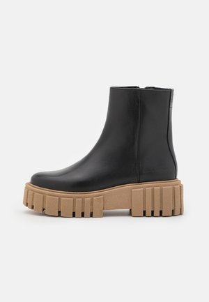 Bota de cuero negra hasta el tobillo con suela plataforma gruesa color beige, que presenta profundos tacones rectangulares y cierre lateral con cremallera.