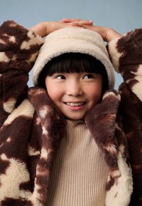 Brązowo-kremowy płaszcz z futerka sztucznego z motywem krowy, w komplecie z kremowym kapeluszem typu bucket. Pod spodem teksturowany sweter w neutralnym odcieniu.