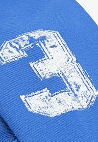 Tessuto blu con un grande numero "3" bianco, consumato, stampato in modo prominente. La texture è liscia con un motivo a maglia.