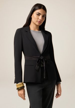 Giovane donna che indossa un blazer nero con cintura legata in vita, top grigio con piccole decorazioni, pantaloni neri e bracciali vistosi al polso sinistro.