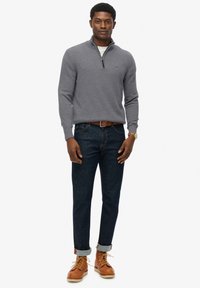 Uomo che indossa un maglione grigio a mezza zip, jeans blu scuro con risvolto, cintura marrone, stivali di pelle beige e orologio d'oro, in piedi contro uno sfondo bianco.