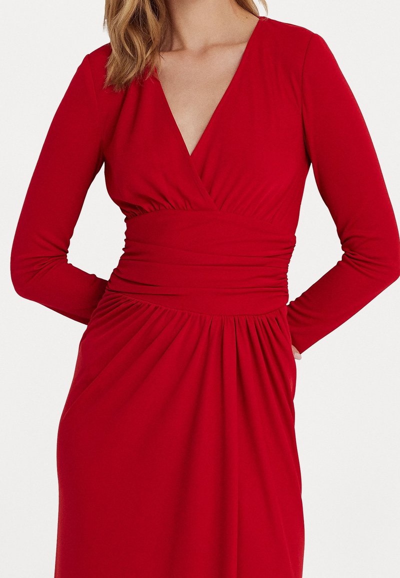 Abito lungo rosso con maniche a lunghezza intera, scollo a V, corpino avvolgente, vita arricciata e gonna fluente. Realizzato in tessuto liscio ed elastico.