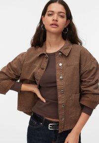 Καφέ denim μπουφάν με γιακά, μπροστινά κουμπιά τύπου σναπ και πλαϊνές τσέπες. Φοράται πάνω από ένα καφέ εφαρμοστό τοπ, συνοδευόμενο από σκούρο τζιν.