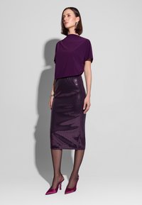 Fioletowa bluzka z krótkim rękawem zestawiona z lśniącą fioletową spódnicą do kolan z cekinami. Modelka ma na sobie fuksjowe szpilki i bransoletkę. Gładka faktura tkaniny.