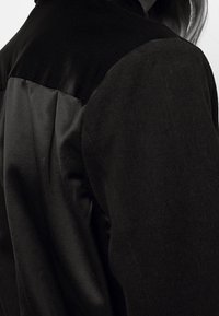 Svart jacka med en blandning av texturerad tyg och släta detaljer, med en skräddarsydd passform som framhäver rynkor på axlarna och ryggen.