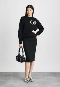 Fekete kötött pulóver fehér "Off" felirattal, testhezálló fekete ceruzaszoknya, szegecses fekete kézitáska és szürke masnival díszített cipők. Semleges háttér.