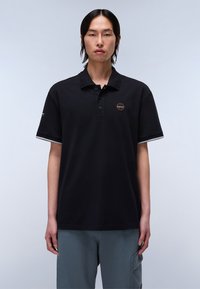 Polo shirt nera con un collettino a tre bottoni, maniche corte con accenti grigi e un logo circolare sul lato sinistro del petto. Texture del tessuto liscia.