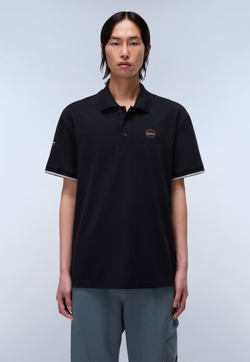 Polo shirt nera con un collettino a tre bottoni, maniche corte con accenti grigi e un logo circolare sul lato sinistro del petto. Texture del tessuto liscia.