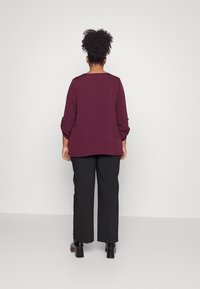 Haut bordeaux à manches longues avec manches retroussées, associé à un pantalon noir ample et des bottes à talons épais noires, vu de dos.