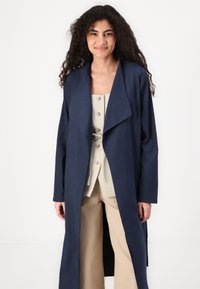 Navy long coat with a shawl collar, open front design, and long sleeves, worn over a beige top with buttons and tied waist detail.