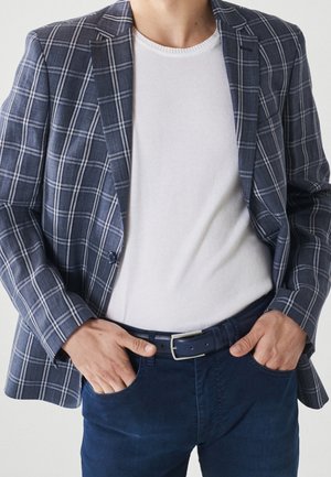 Homme portant un t-shirt blanc à col rond, un blazer à carreaux bleu marine, un pantalon bleu foncé et une ceinture, les mains dans les poches, devant un fond uni.