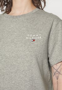 Grå t-shirt med kort ärm i bomull, med "TOMMY HILFIGER"-logotypen i vitt ovanför en liten flaggrafik på vänster bröst.