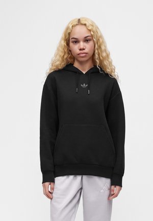 Ung person med langt, krøllet blondt hår iført en sort Adidas hættetrøje og lysegrå Adidas bukser, stående mod en ensfarvet baggrund.