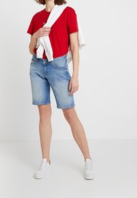 En kvinna står med en vit tröja hängande över axeln, iförd en röd t-shirt, denim shorts och vita sneakers mot en enfärgad bakgrund.