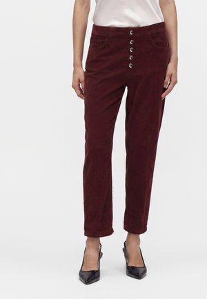 Femme portant un pantalon bordeaux à boutons sur le devant et des talons noirs à bout pointu, debout devant un fond clair uni.
