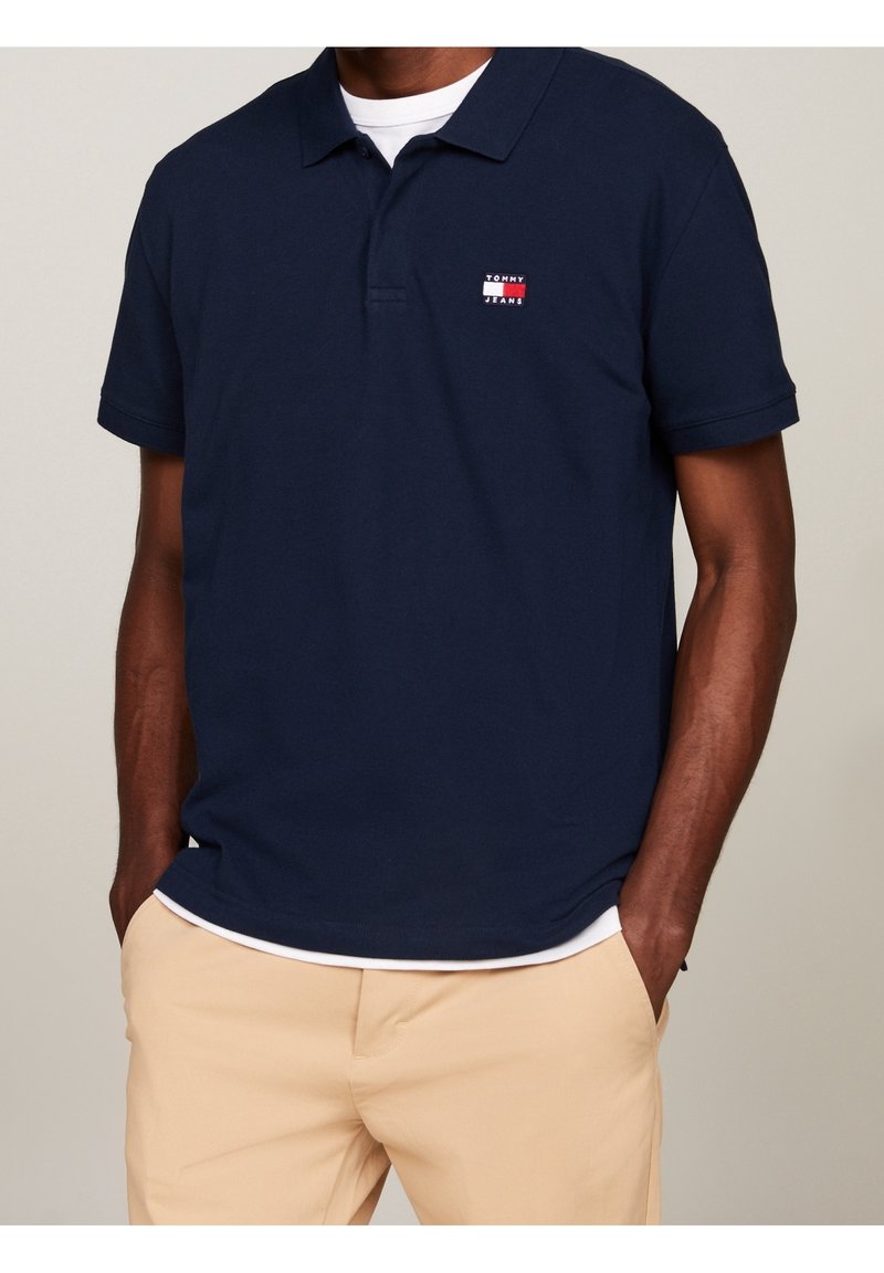 Homme portant un polo bleu marine Tommy Jeans par-dessus un t-shirt blanc et un pantalon beige, les mains dans les poches, sur un fond uni.
