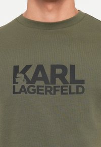 Olīvzaļš kokvilnas T krekls ar izteiksmīgu melnu "KARL LAGERFELD" logotipu un siluetu grafiku, apaļu kakla izgriezumu un īsām piedurknēm.