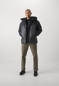 Puffer jacket in black, featuring a high collar and zip closure. Paired with olive-green cargo pants and black sneakers. Minimalist design.