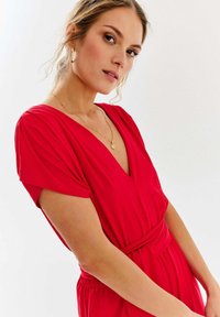 Czerwona kombinezon z krótkimi rękawami, głębokim dekoltem w serek, paskiem w talii i zwiewnym materiałem. Gładka faktura i dopasowany krój. Widoczna złota naszyjnik.