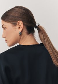 Silberne ovale Ohrringe, ein schwarzes Haargummi mit einem Kristallakzent und ein schwarzes Oberteil. Das Haar ist zu einem glatten Pferdeschwanz zurückgebunden und zeigt das Profil.