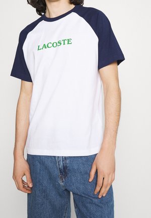 Osoba nosząca białą koszulkę z krótkim rękawem z granatowymi rękawami i zielonym napisem "LACOSTE", połączoną z niebieskimi jeansami, na jednolitym tle.