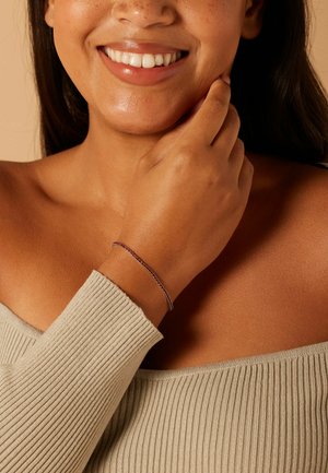 Bracelet fin violet porté au poignet, présentant un design délicat à chaîne. La personne a la peau olive et porte un haut gris clair côtelé.