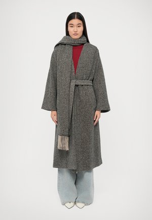 Manteau long en chevrons gris avec des manches larges et une écharpe assortie à bords frangés, superposé sur un top rouge et un jean bleu clair.
